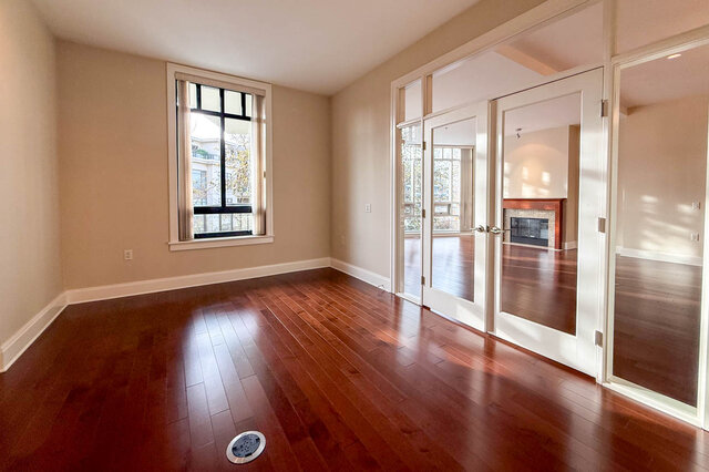 Spacious living room with hardwood floors, large window, and sliding glass doors leading to a cozy fireplace. - 301-605 Clyde Ave, West Vancouver