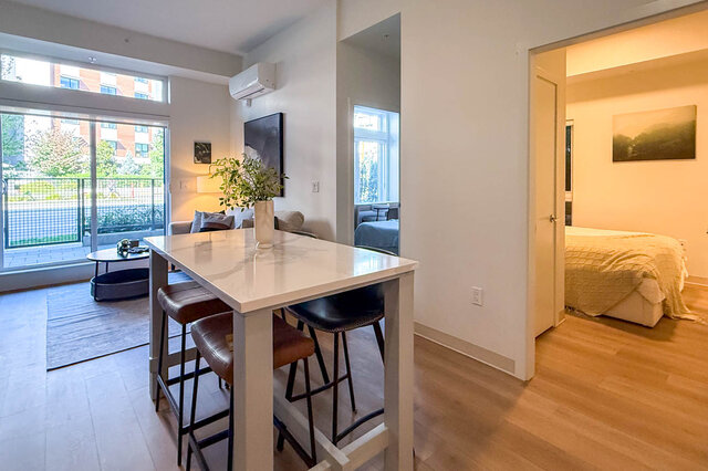Dining area with a white marble table, leather bar stools, and hardwood floors opening to a bedroom. - 115-1529 Oxford Street, North Vancouver