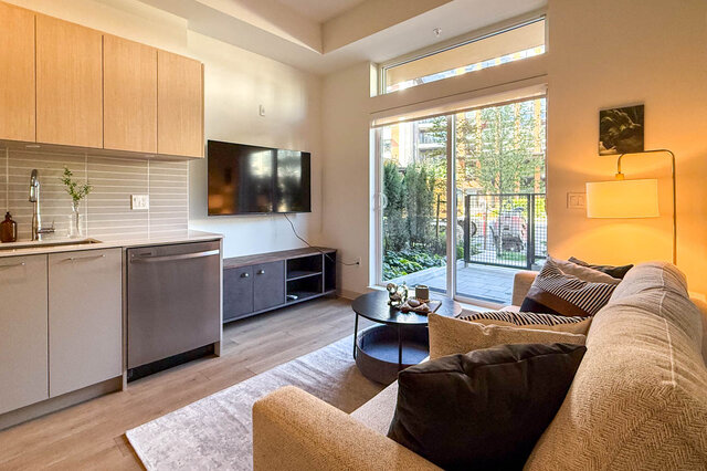 Open-concept living room with hardwood floors, kitchenette, and large window to a balcony. - 115-1529 Oxford Street, North Vancouver