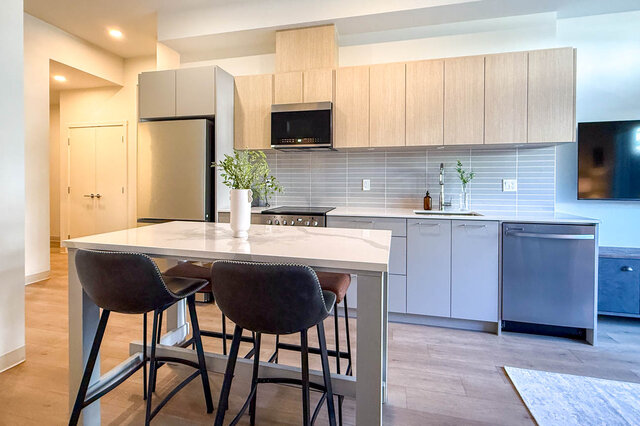 Modern kitchen with light wood cabinetry, white marble island, and stainless steel appliances over hardwood floors.