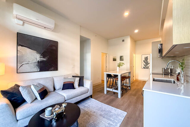 Open-concept living room with light gray sofa, hardwood floors, and adjacent white kitchen with stainless sink.