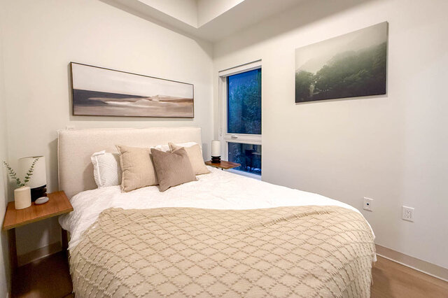 Neat bedroom with a neutral bed, wood nightstands, and framed landscape art on white walls. - 115-1529 Oxford Street, North Vancouver