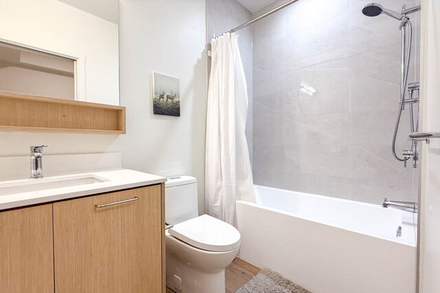 Clean bathroom with a light wood vanity, white countertop, and a tub-shower combo with gray tile walls. - 115-1529 Oxford Street, North Vancouver