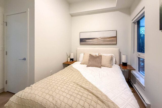 Neutrally furnished bedroom with upholstered headboard, wooden nightstands, and a window overlooking trees. - 115-1529 Oxford Street, North Vancouver