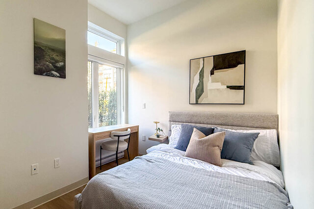 Bright bedroom with hardwood floors, a window desk nook, and a bed dressed in gray linens with blue and tan pillows.