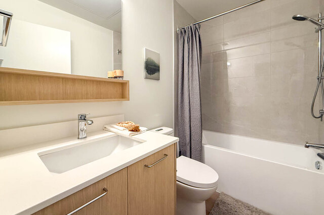 Modern bathroom with a white countertop, light wood vanity, and a tub-shower combo with gray tile. - 115-1529 Oxford Street, North Vancouver