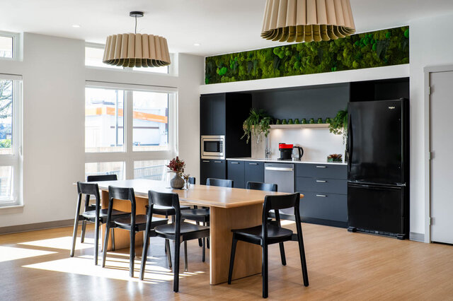 Dining area with light wood table, dark grey cabinetry, and a living moss wall accent above the kitchen. - 115-1529 Oxford Street, North Vancouver