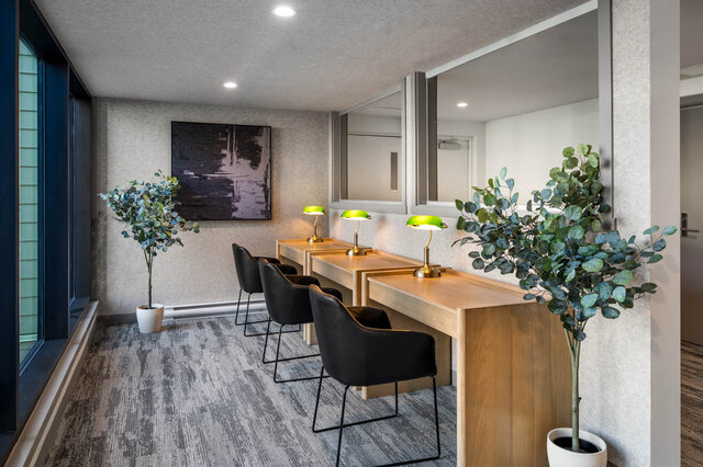 Vanity area with three wooden desks, green-shaded lamps, black chairs, and potted eucalyptus plants. - 115-1529 Oxford Street, North Vancouver