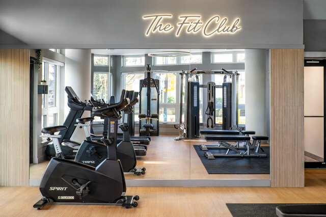Modern fitness room with hardwood floors, cardio bikes, and weight machines under a glowing "The Fit Club" sign.