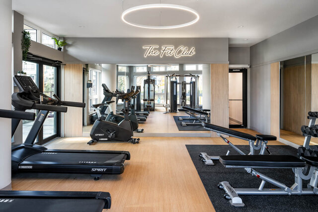 Modern fitness room with hardwood floors, treadmills, and weight benches beneath a circular ceiling light. - 115-1529 Oxford Street, North Vancouver