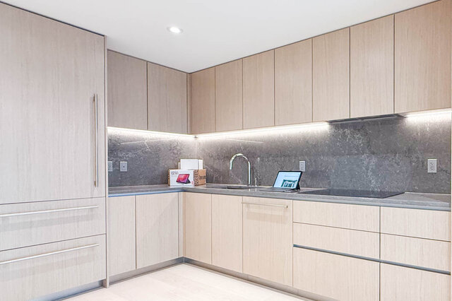 This is a kitchen with light wood cabinets, under-cabinet lighting, a stainless steel faucet, and a gray countertop.