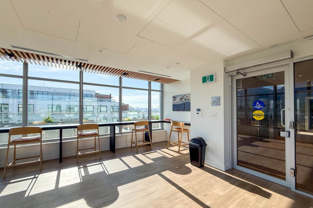 The lobby has floor-to-ceiling windows, bar stools, a trash can, and glass entrance doors. - 406-777 Harbour Way, North Vancouver
