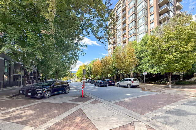 The building exterior shows a street with parked cars, mature trees lining both sides, and a tall residential building in the