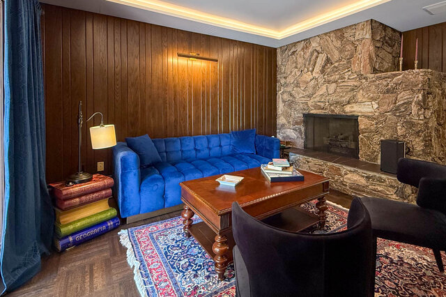 A well-lit living room featuring a blue velvet sofa, stone fireplace, and wooden panel walls. - 1249 Chartwell Place, West Vancouver