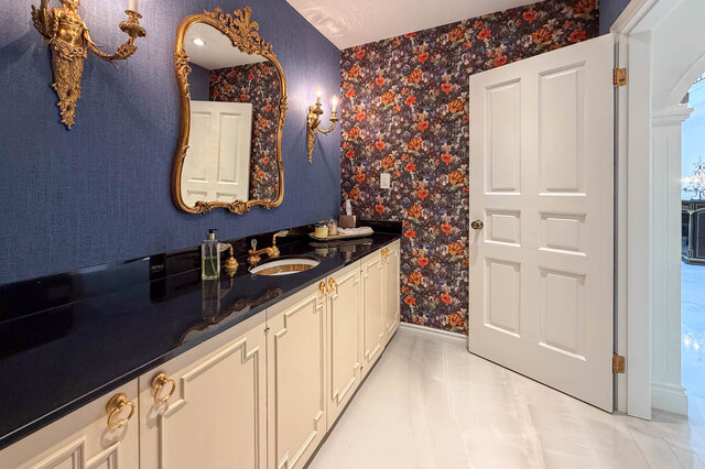 A well-lit bathroom with gold fixtures, floral wallpaper, and cream cabinets - 1249 Chartwell Place, West Vancouver