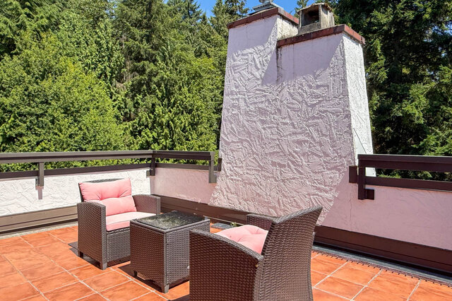 Balcony with wicker chairs, pink cushions, terracotta tiles, and a fireplace structure against a forest backdrop.