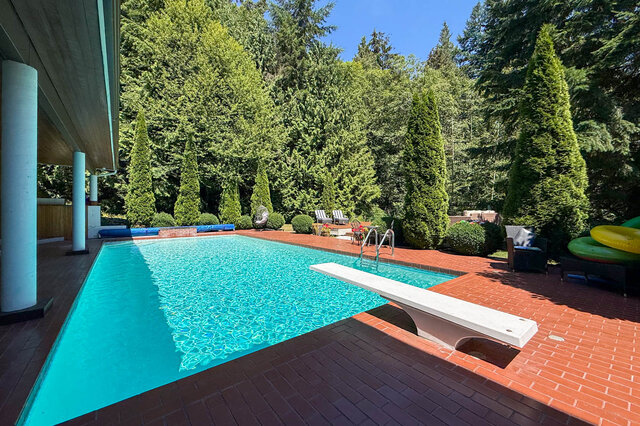 Sunny outdoor pool area with brick patio, lounge chairs, and surrounding evergreen trees - 1249 Chartwell Place, West Vancouver