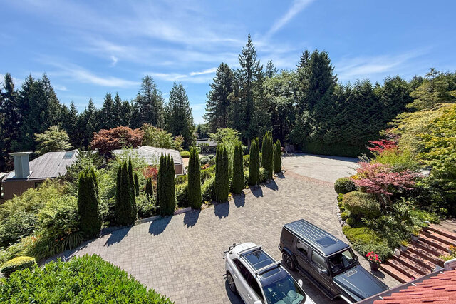Aerial view of a spacious property with a large driveway, lush landscaping, and surrounding forest under a bright blue sky