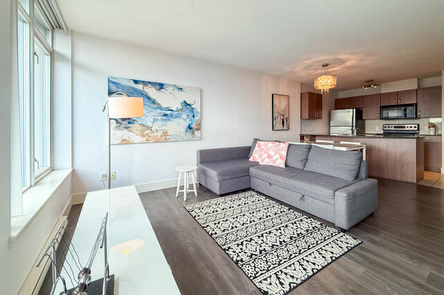 Bright living room with gray sectional sofa and modern kitchen, featuring hardwood floors and large windows.