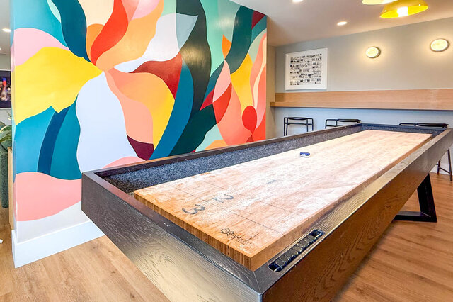 Game room with vibrant abstract mural wall and wooden shuffleboard table on hardwood floors. - 204-1680 East 12th Avenue, Vancouver East Side