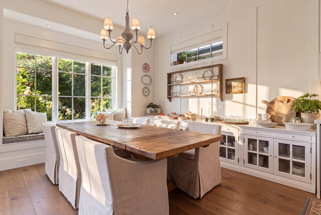 A bright, spacious dining area with a wooden table, white upholstered chairs, and large windows overlooking greenery