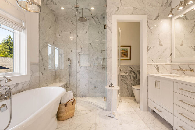 A bright, marble-clad bathroom with a freestanding tub, glass shower, and window view - 1248 Kings Avenue, West Vancouver