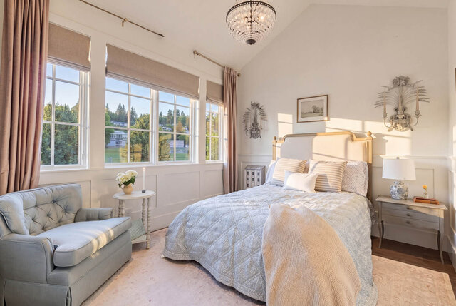 A bright, spacious bedroom with large windows overlooking greenery, featuring a bed, chandelier, and upholstered sofa