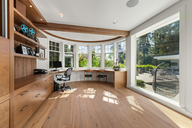 The den features a curved desk, office chair, wooden built-in shelves and cabinets, and large windows overlooking a garden.