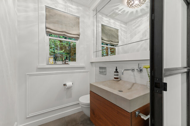 This bathroom has a concrete sink countertop, a toilet, a window with a Roman shade, and wainscoting. - 2940 Mathers Avenue, West Vancouver