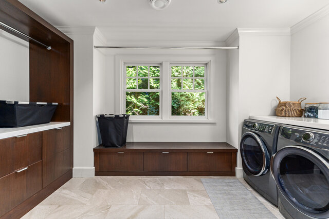 This is a laundry room with stainless steel appliances, dark wood cabinetry, a window with a green view, and laundry storage.