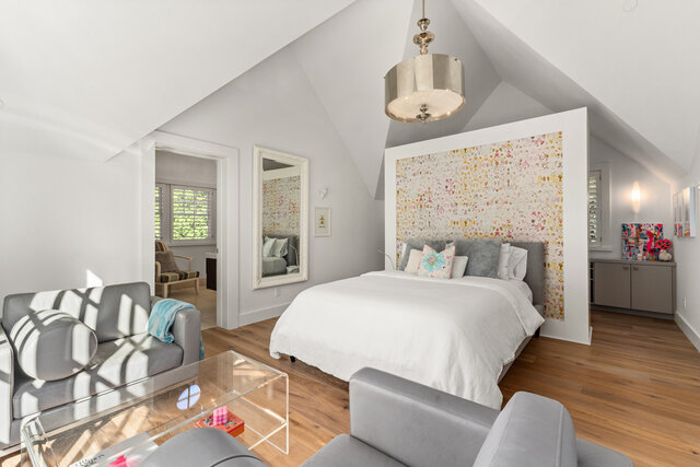 This is a bedroom with a bed, gray sofa, glass coffee table, large mirror, chandelier, and hardwood floors. - 2940 Mathers Avenue, West Vancouver