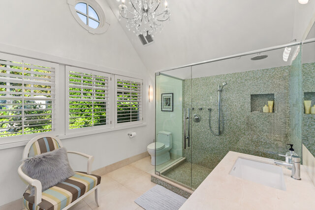This bathroom features a glass-enclosed shower, a toilet, a sink with a countertop, a crystal chandelier, windows with white 