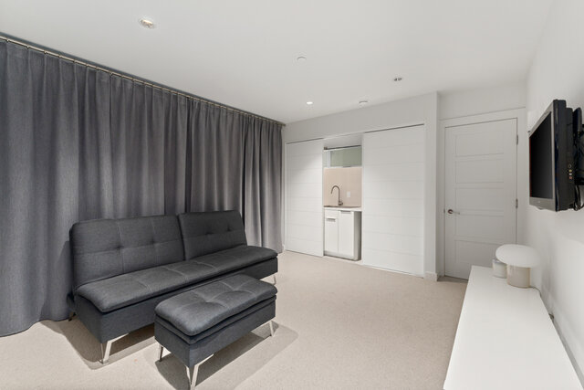 The living room contains a gray sofa, a footrest, a wall-mounted TV, gray curtains, and a carpeted floor. - 2940 Mathers Avenue, West Vancouver