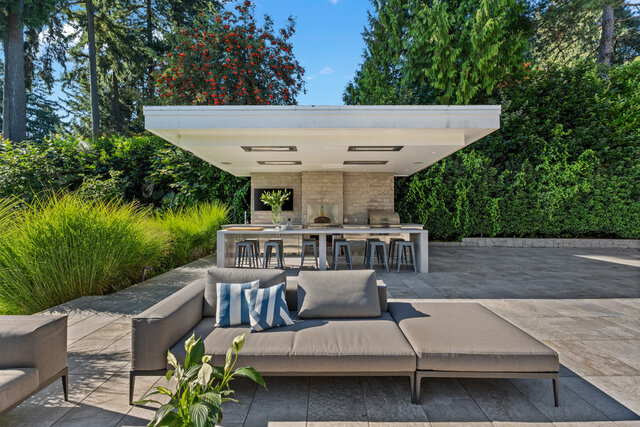 The lounge has a sofa with striped cushions, a covered patio with a fireplace, and is surrounded by mature trees.