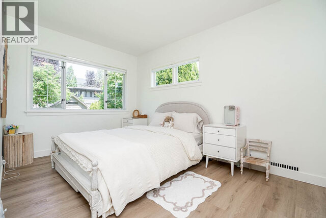 A bedroom with a bed, two nightstands, a chair, a small table, and multiple windows showing greenery outside