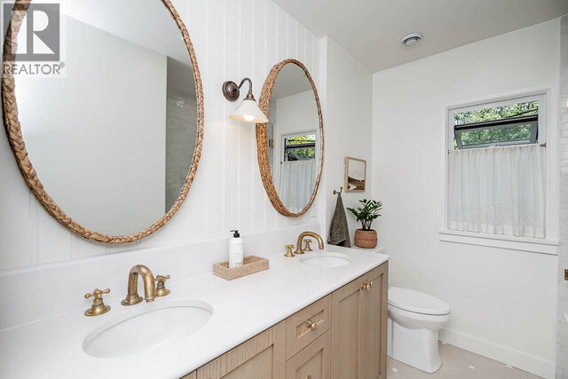 This is a bathroom with a double vanity, two round woven mirrors, brass faucets, a toilet, a window with a curtain, and a pot