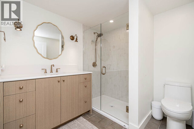 The bathroom features a light wood vanity with a white countertop, a round mirror, a glass-enclosed shower, and a white toile