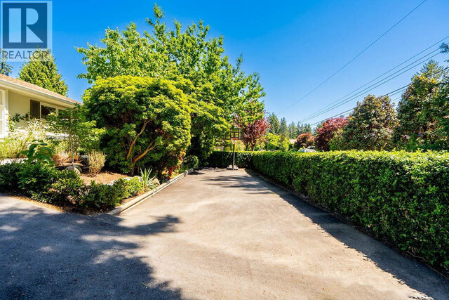 The building exterior features an asphalt driveway, neatly trimmed hedges, mature trees, and landscaped garden areas.