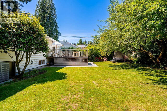 The building exterior features a deck with railing, a patio umbrella, a green lawn, and mature trees surrounding the property