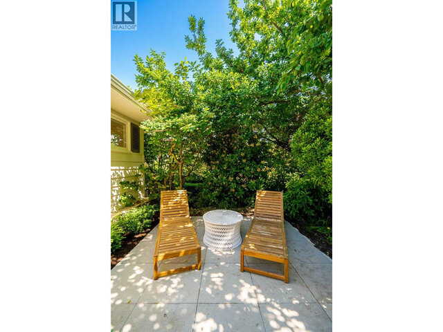 The lounge area has two wooden lounge chairs, a wicker side table, and is surrounded by mature trees on a concrete patio.