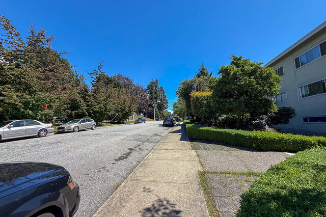 The building exterior with a paved sidewalk, parked cars, and mature trees along the street - 102-461 16th West street, North Vancouver