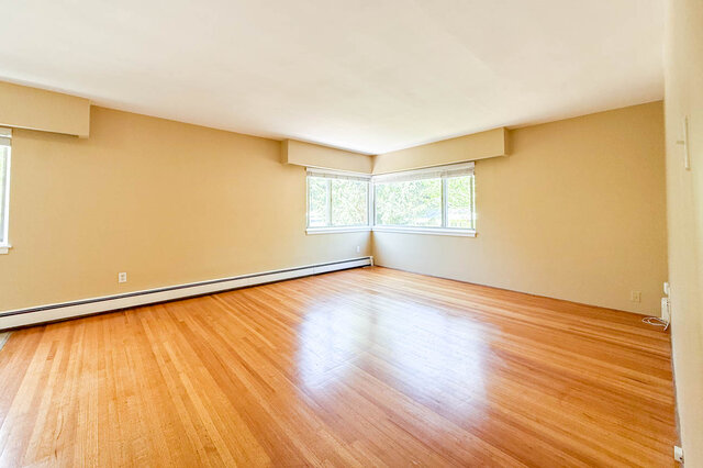 This is a living room with hardwood floors, beige walls, baseboard heating, and windows. - 102-461 16th West street, North Vancouver