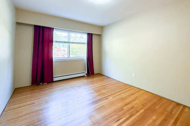 This is a bedroom with hardwood floors, a window with purple curtains, and an electrical outlet on the wall.