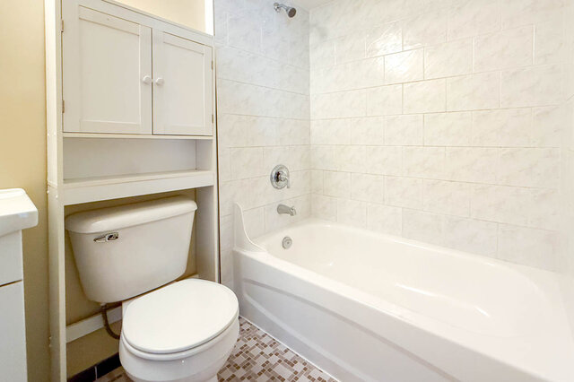 This bathroom contains a white toilet, a bathtub with a showerhead, a cabinet with storage above the toilet, and a small wind