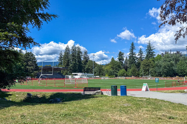 The view shows a sports field with a running track, soccer goals, tall trees, and a clear blue sky. - 102-461 16th West street, North Vancouver