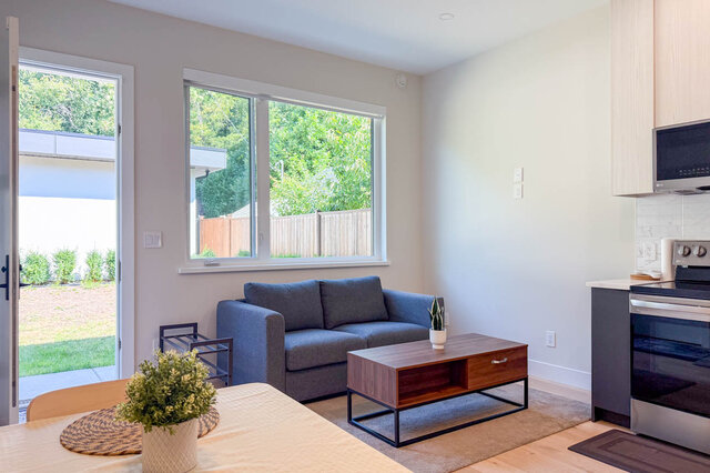 Furnished living room with gray sofa, wood coffee table, and large windows overlooking greenery. - Suite-1384 Hope Road, North Vancouver