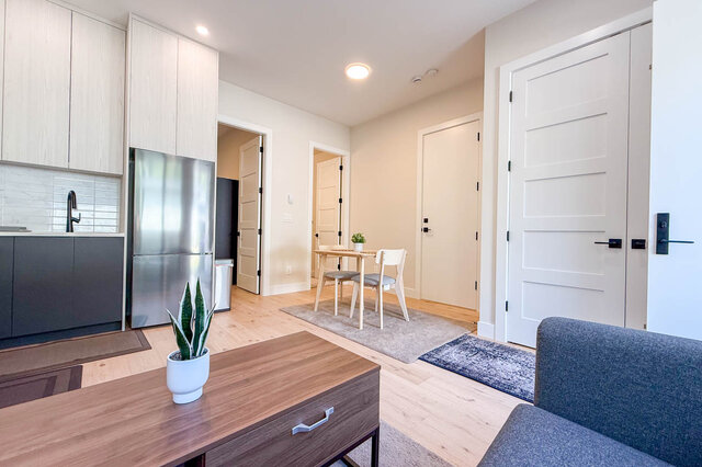 Furnished open living area with light hardwood floors, stainless steel fridge, and a small dining nook. - Suite-1384 Hope Road, North Vancouver