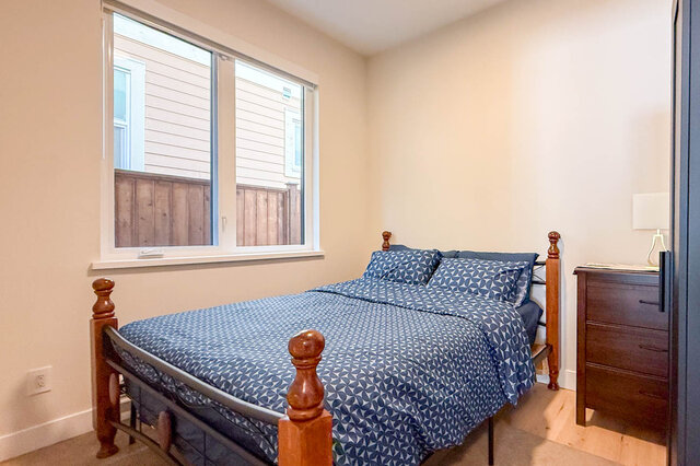 Furnished bedroom with hardwood floors, a metal-and-wood bed, and a window overlooking a backyard fence. - Suite-1384 Hope Road, North Vancouver