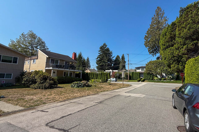 Quiet street view of a two-story home with a balcony, manicured hedges, and tall evergreens under a clear blue sky.