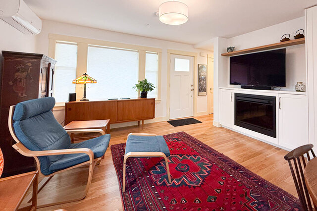 Furnished living room with hardwood floors, a red patterned rug, and a blue lounge chair beside a built-in fireplace.